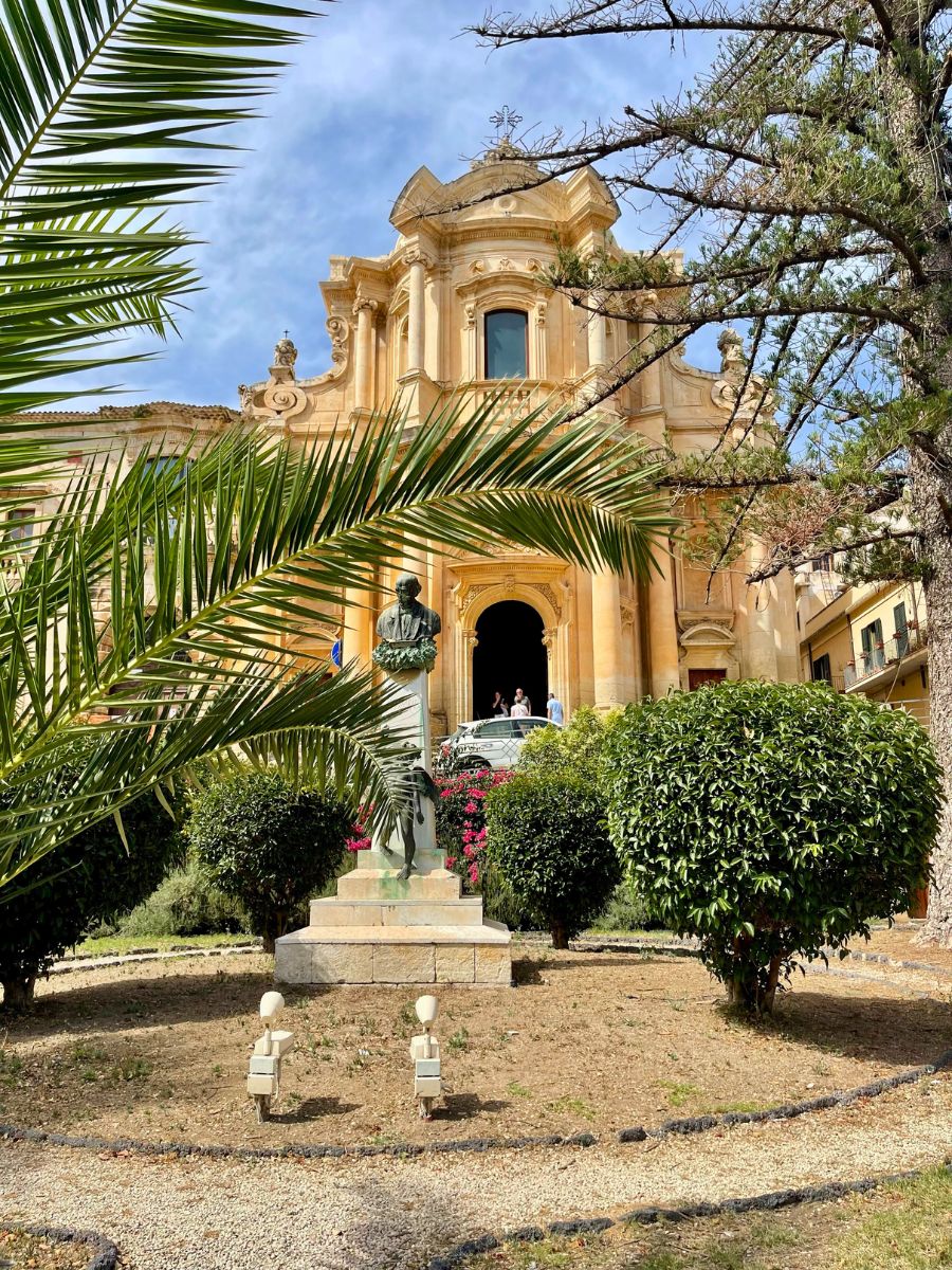 A lush garden scene in Noto, Sicily, featuring a statue of a man standing on a stone pedestal in the center. The statue is surrounded by neatly trimmed bushes, palm fronds, and decorative trees. In the background stands a majestic Baroque church with elaborate architectural details, including tall columns, sculpted ornamentation, and arched windows. The warm golden tones of the church’s façade contrast beautifully with the greenery in the foreground and the soft blue sky above.