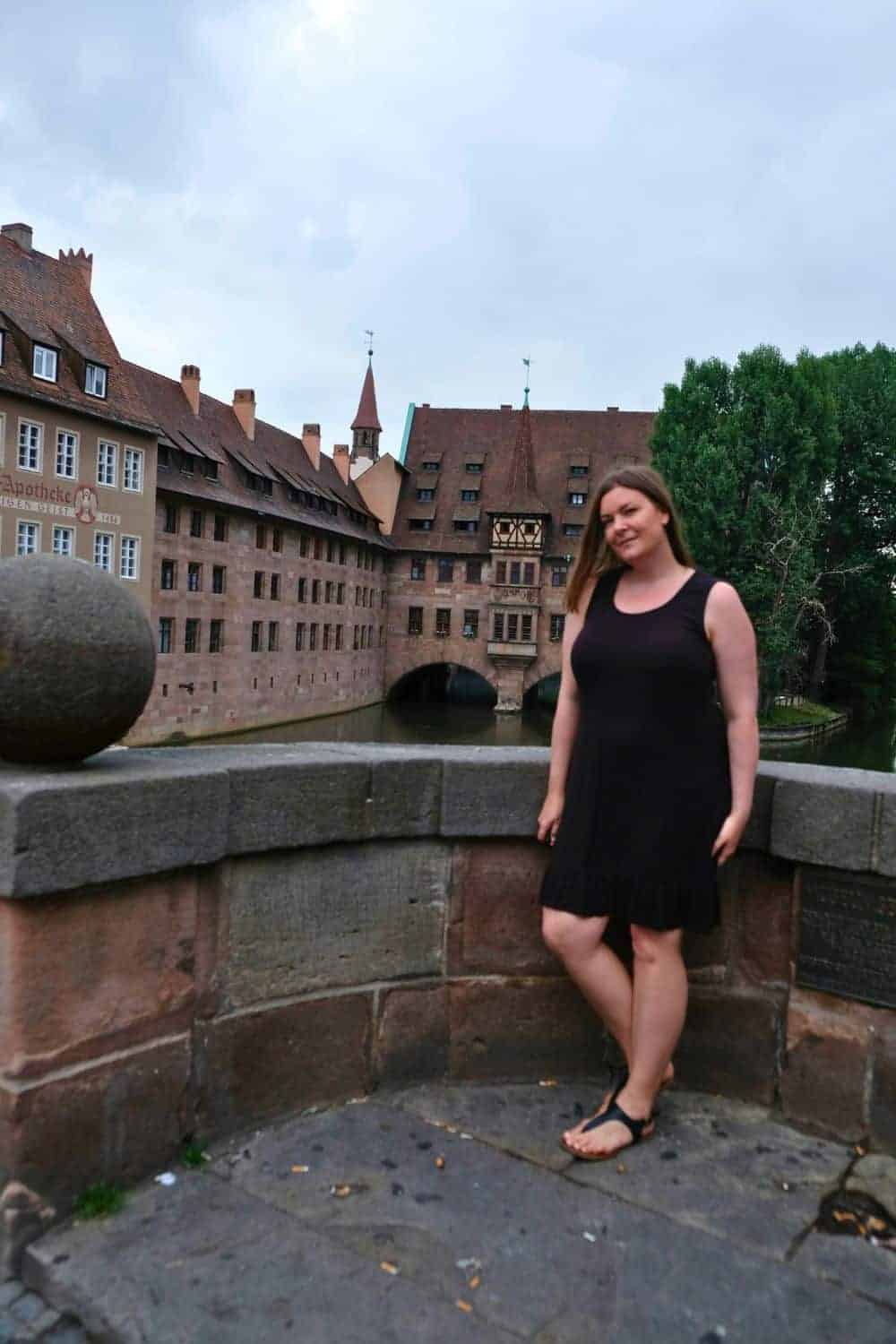 A solo woman traveler stands on an old bridge, with historic sandstone buildings and a peek of the Pegnitz River in the background. The overcast sky casts a soft light on the scene, enhancing the timeless beauty of the city.
