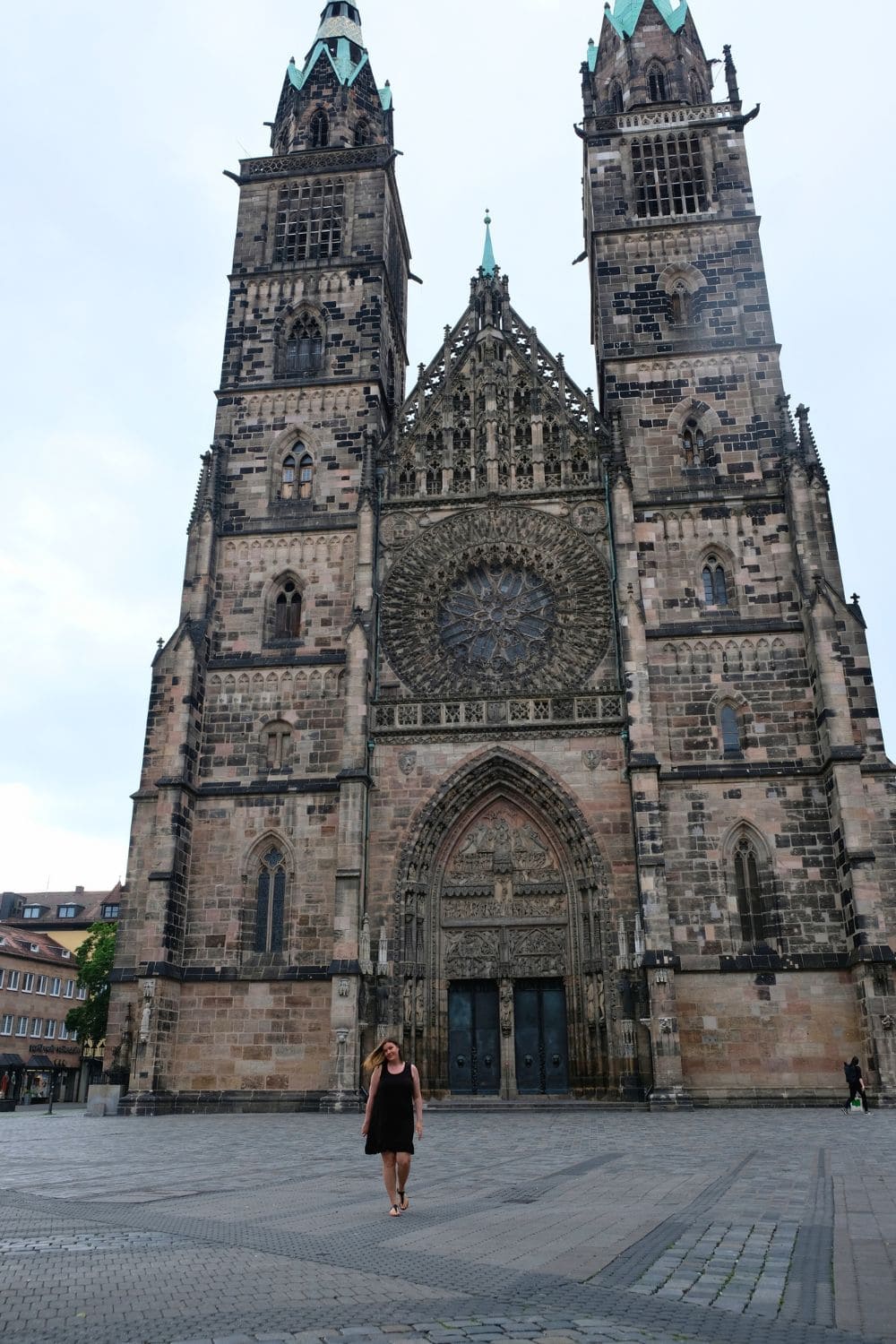 A woman in front of the magnificent Gothic facade of Nuremberg's St. Lorenz Church. The church's intricate stonework, grand spire, and ornate rose window are highlighted by the sun, showcasing this architectural marvel. 