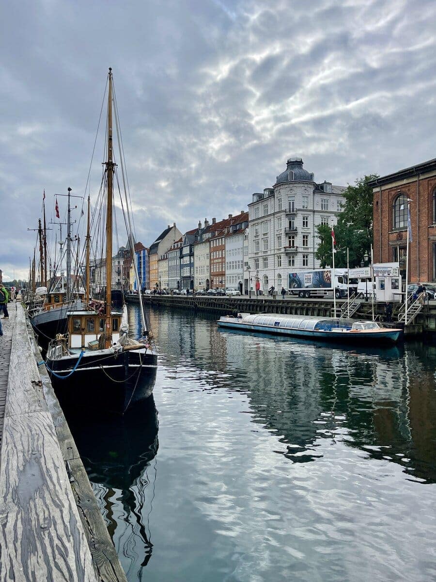 A scenic view of Copenhagen’s canal with historic sailboats moored along the waterfront, complemented by traditional architecture under a dramatic cloud-filled sky.