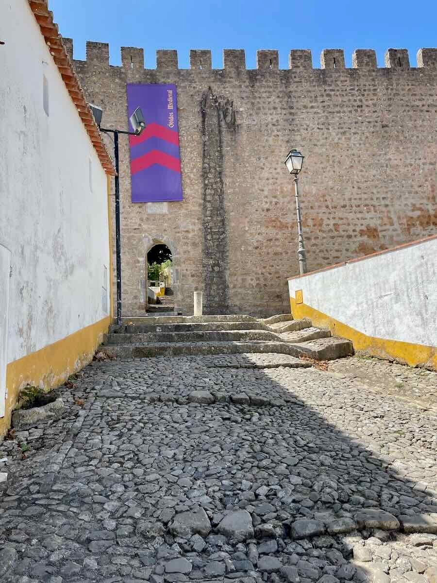 The cobblestone path leading to the castle walls of Óbidos, Portugal. A small archway and banner can be seen on the ancient stone walls, reflecting the town’s medieval charm.