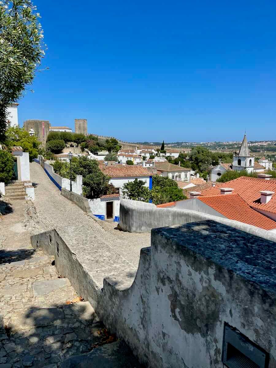 A panoramic view of Óbidos, Portugal, with its red-tiled rooftops, historic church, and the surrounding lush countryside under a clear blue sky.