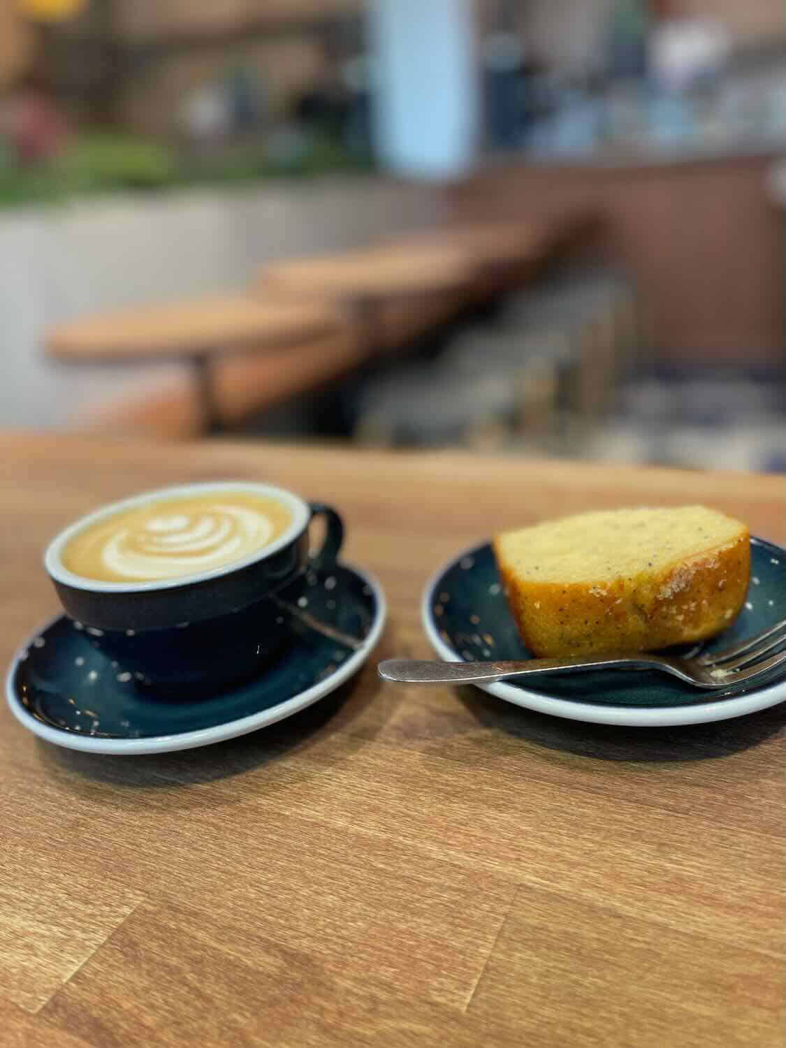 A close-up image of a cozy moment at a Paris café, featuring a cup of cappuccino with artful froth and a slice of lemon cake on dark blue ceramic dishes. The warm, inviting ambiance suggests a perfect break for any traveler needing a moment of relaxation.