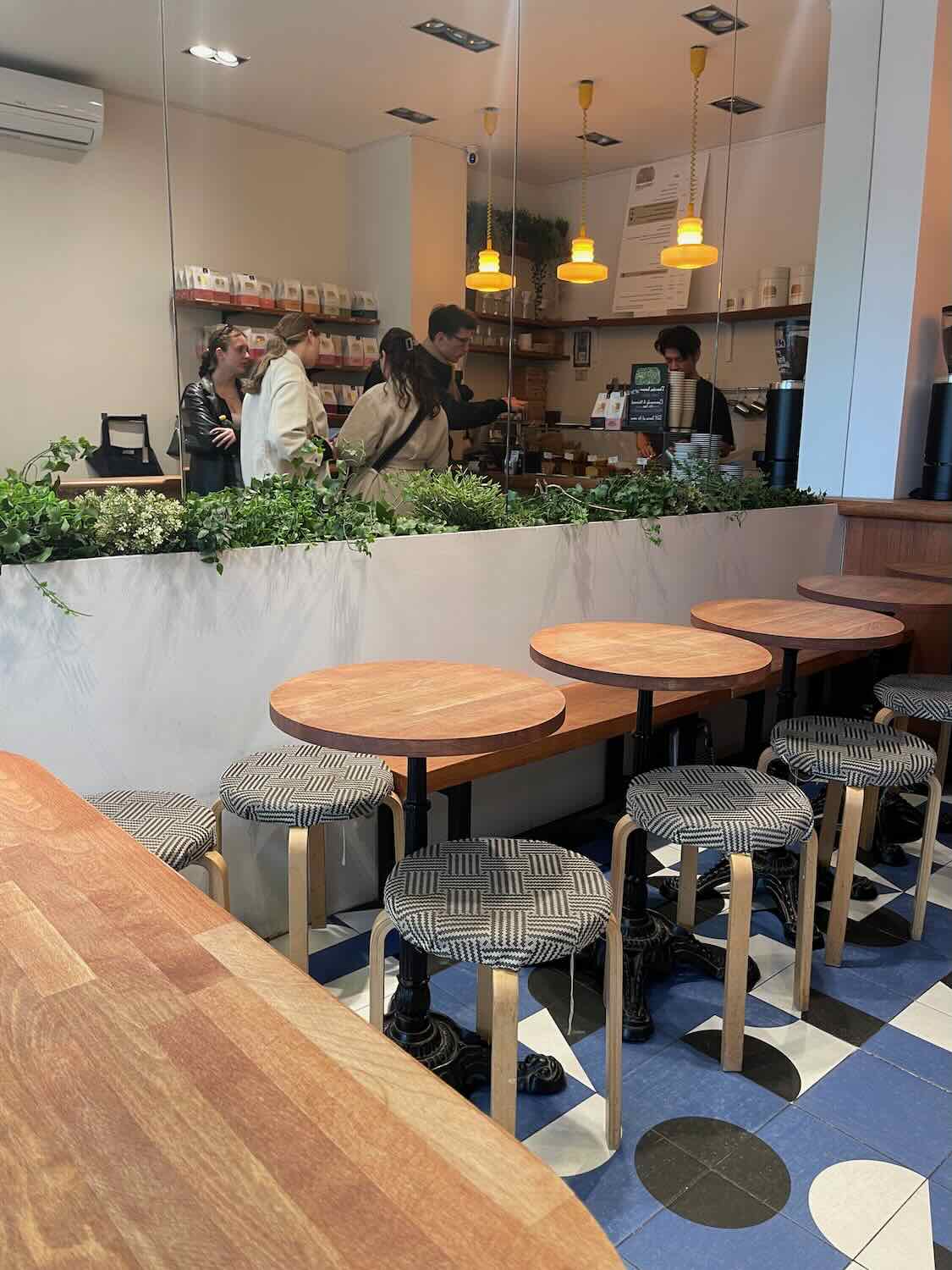 An interior view of a bustling Paris café, showcasing a modern and inviting space. Wooden tables and chairs with patterned cushions are arranged neatly, while customers engage casually at the counter under warm yellow lights, depicting a lively scene typical of Parisian life.