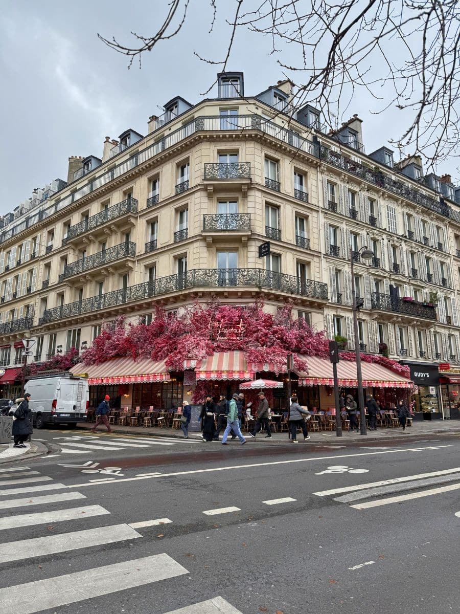 A corner building in Paris with ornate wrought-iron balconies and a charming café on the ground floor. The café features striped pink-and-white awnings adorned with vibrant pink floral decorations, with people walking by on a cloudy day.