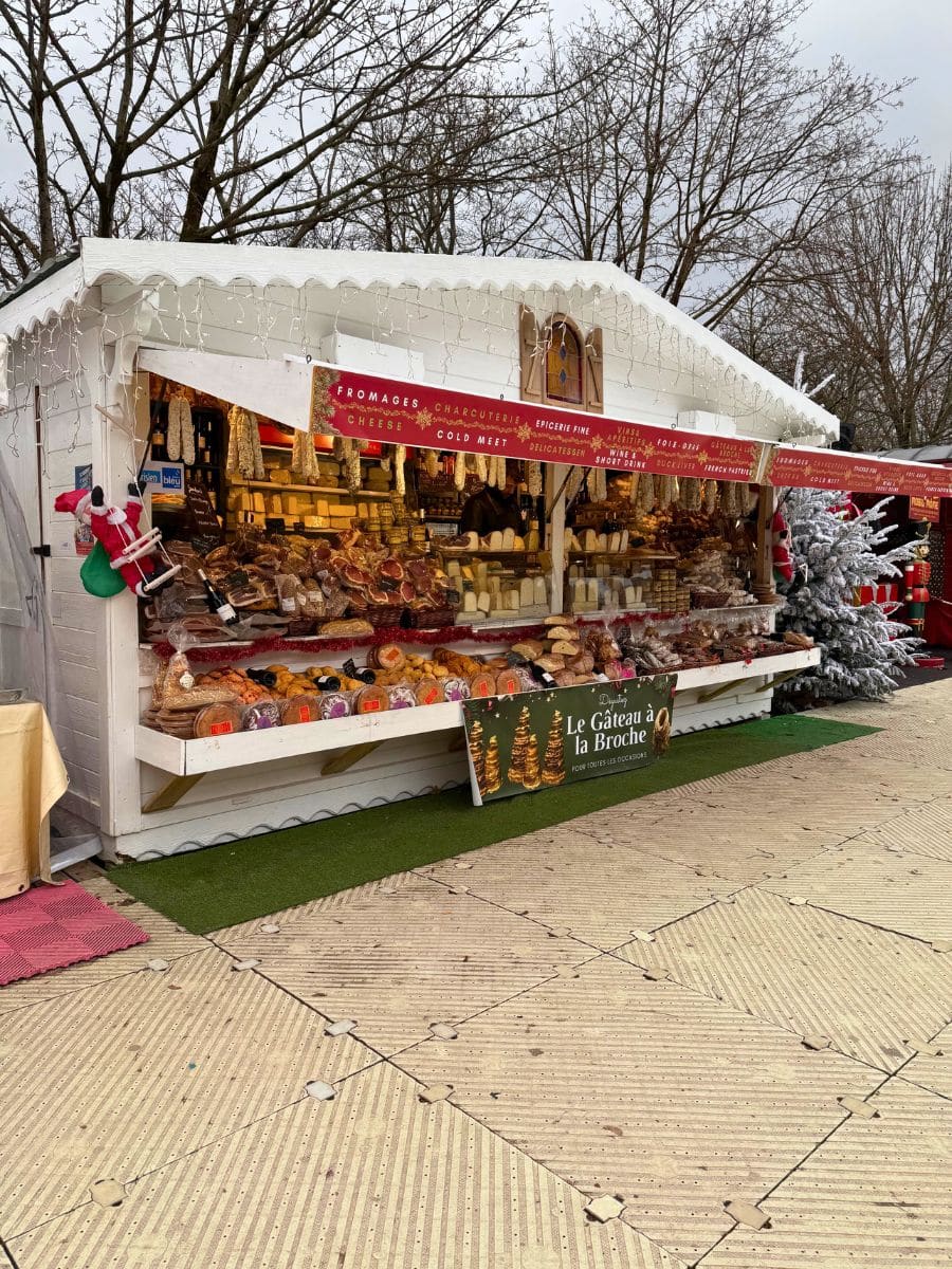 A charming white stall at a Paris Christmas market displaying a variety of French cheeses, cured meats, and other delicacies. The booth is adorned with festive decorations and fairy lights, with a small sign promoting "Le Gâteau à la Broche" in front. Bare trees and holiday-themed stalls are visible in the background.