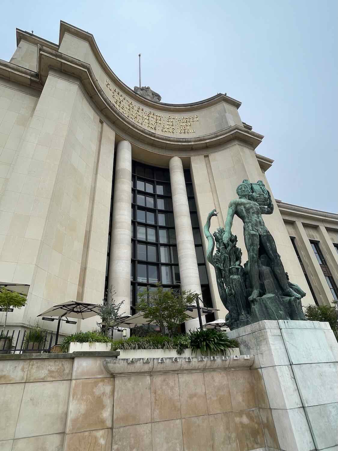 A view of the Palais de Chaillot in Paris, featuring towering columns and statues, including a dramatic sculpture of a man and a horse at the forefront, under an overcast sky.