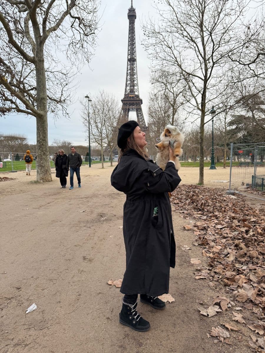 Melissa holding Teddy near the Eiffel Tower in Paris, surrounded by trees and fallen leaves on a cloudy day.