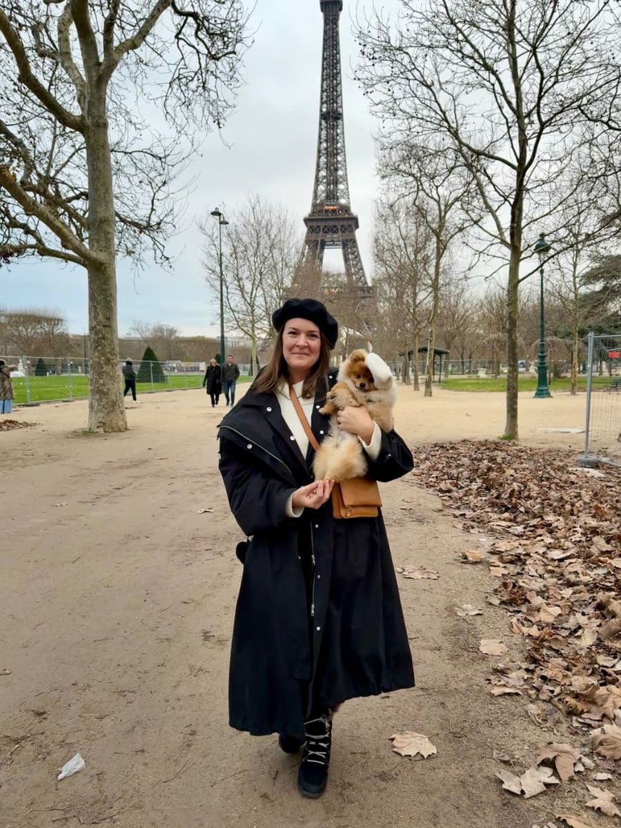 Melissa in a black beret and coat stands in front of the Eiffel Tower holding Teddy, a fluffy Pomeranian dog wearing a white beret, surrounded by bare trees and fallen leaves in a park setting.