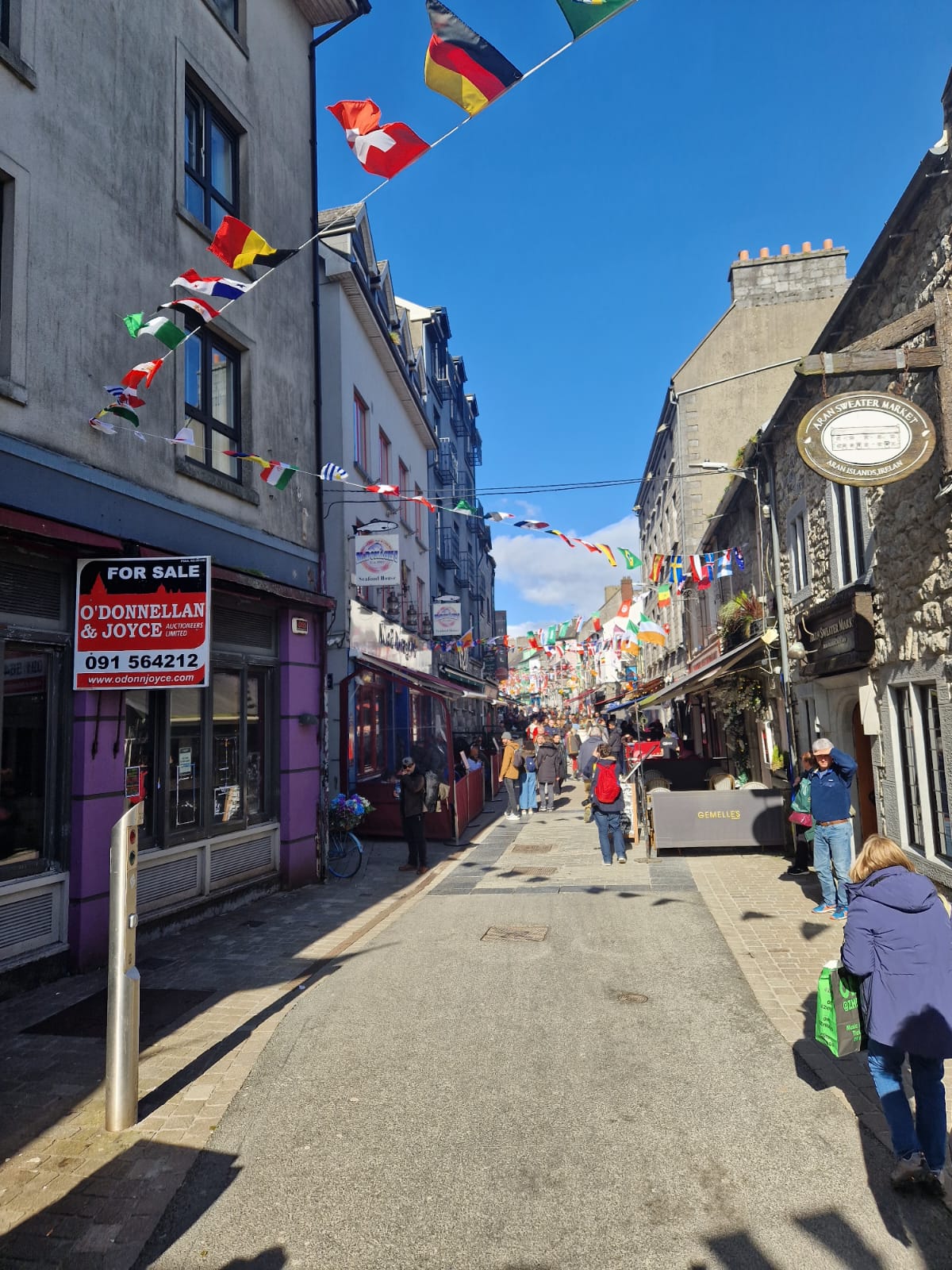 A lively street in Galway, Ireland, with colorful international flags strung across the road. Shops and restaurants line the street, and people stroll along the pedestrian area under a bright blue sky.