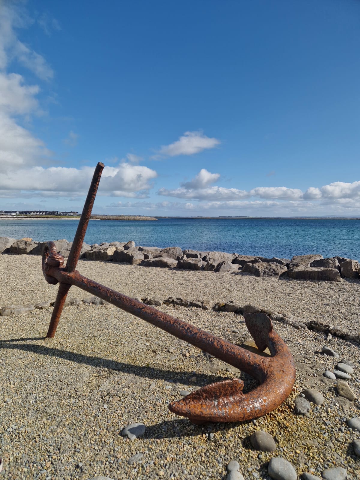 A rusty anchor resting on a gravel beach with a serene view of Galway Bay in the background. The calm blue water meets a clear sky dotted with white clouds, adding to the tranquil coastal scene.