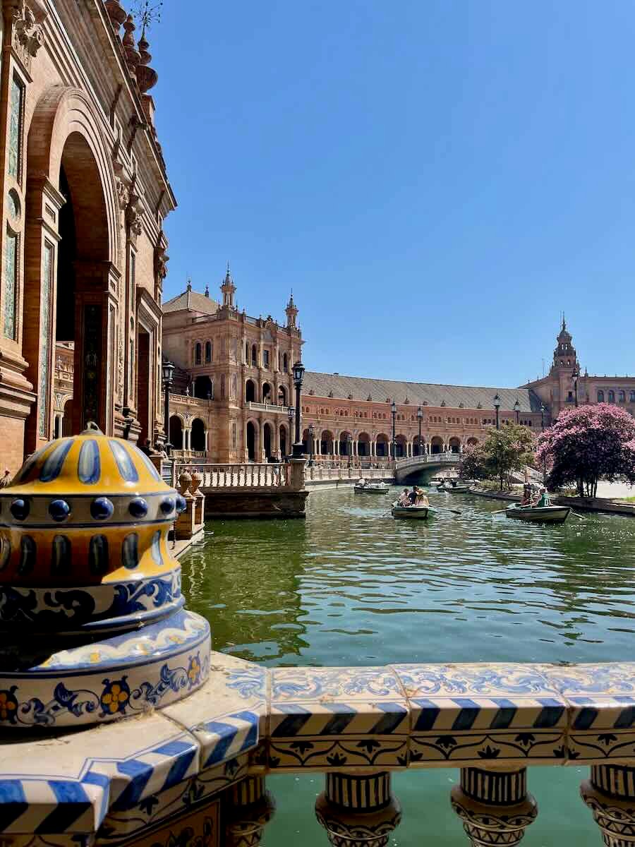 A scenic view of Plaza de España in Seville, Spain. The image captures the beautiful architecture of the semi-circular building, a ceramic balustrade in the foreground, a canal with boats, and blooming trees, all under a bright blue sky.