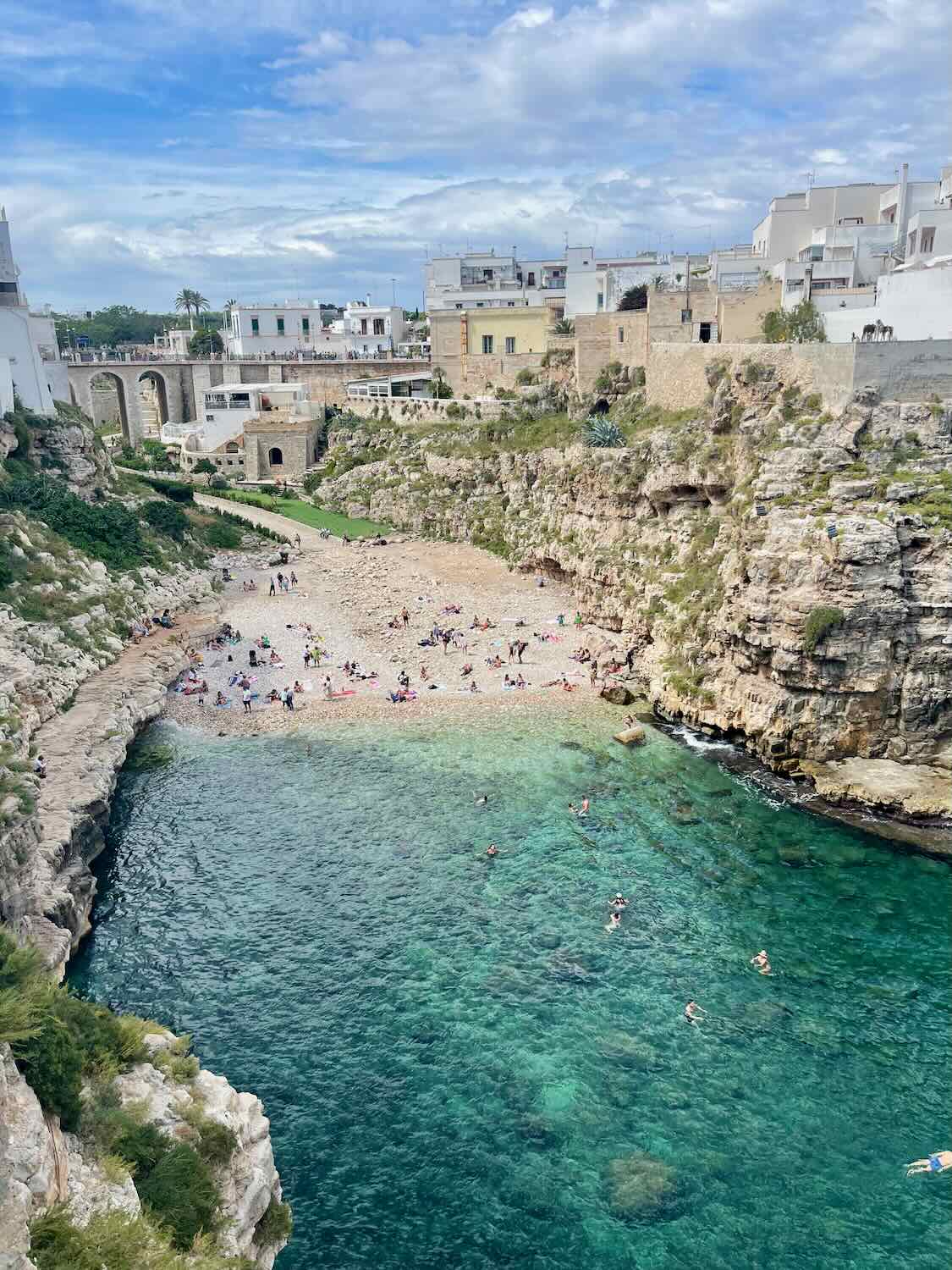 A view of the beach at Polignano a Mare, Italy, with turquoise water and rocky cliffs surrounding the cove. People are sunbathing on the pebble beach and swimming in the clear water. The town with its white buildings is visible in the background.
