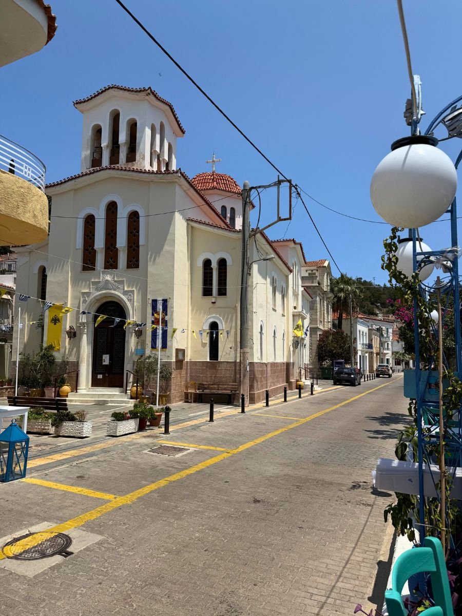 A picturesque Greek Orthodox church with red-tiled domes and festive banners lining a quiet street in Poros, Greece, under a bright blue sky.