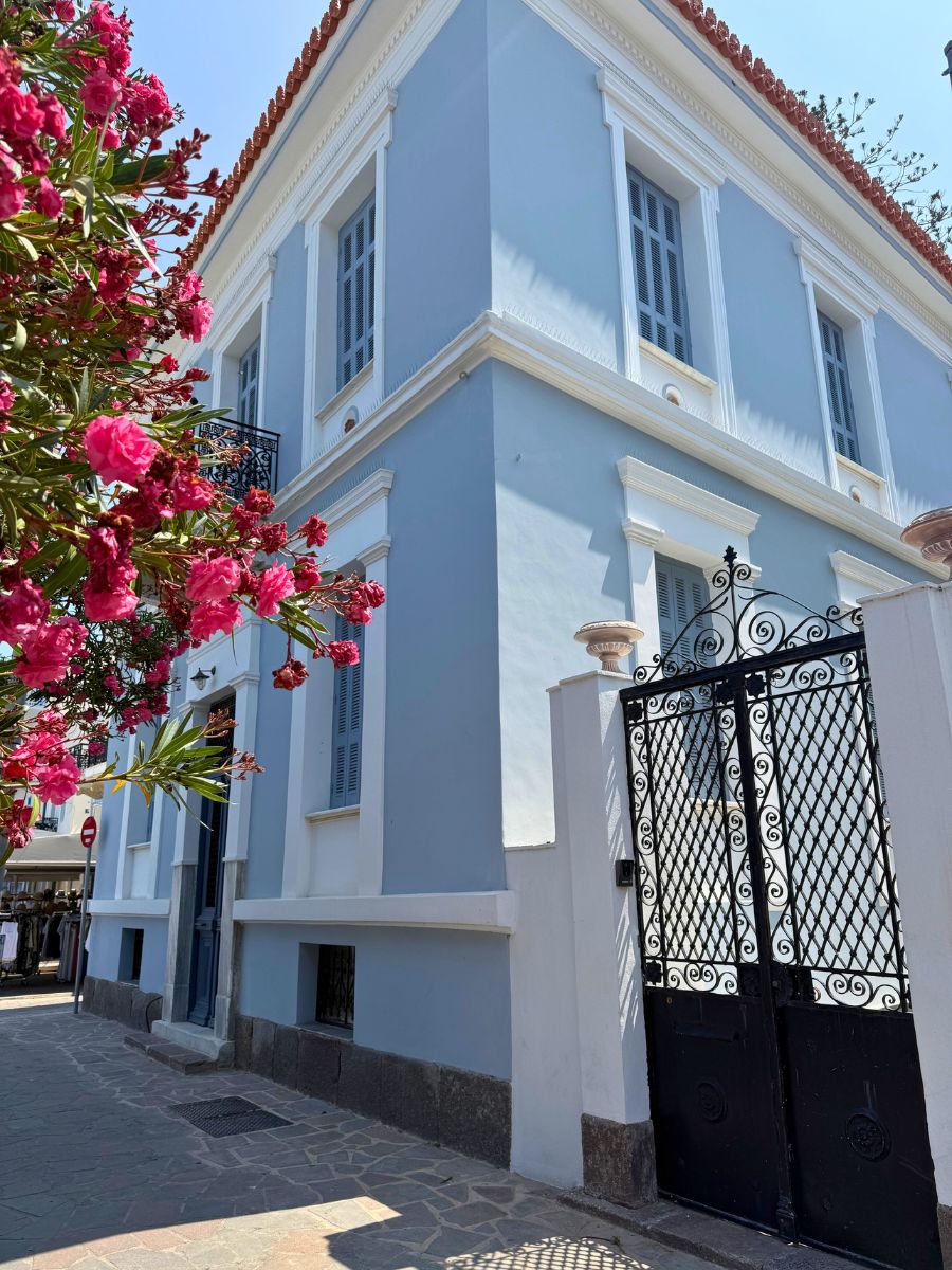 Elegant light blue neoclassical building with white trim and iron gate in Poros, framed by blooming pink flowers on a sunny day.