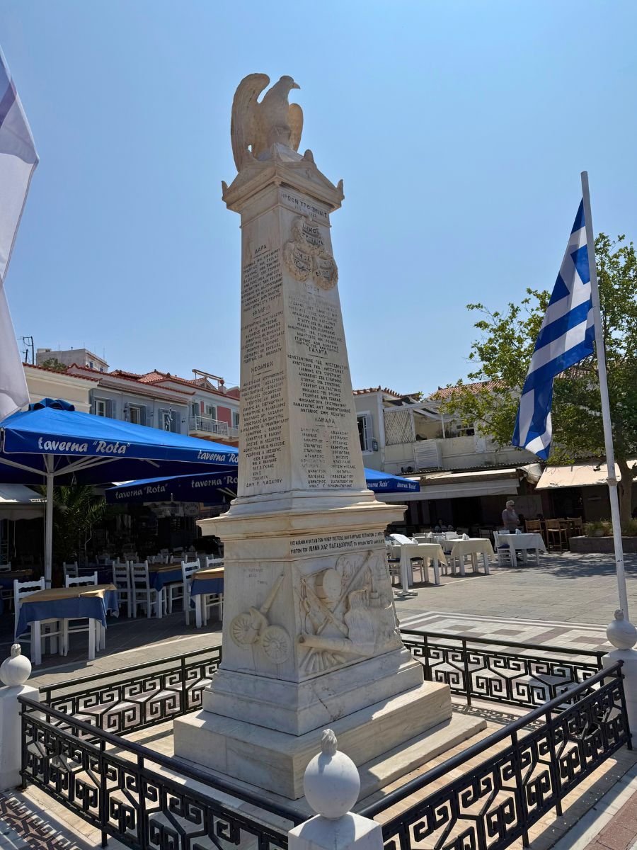 Marble war memorial monument with an eagle sculpture on top, located in a town square in Poros, Greece, surrounded by Greek flags and outdoor tavernas.