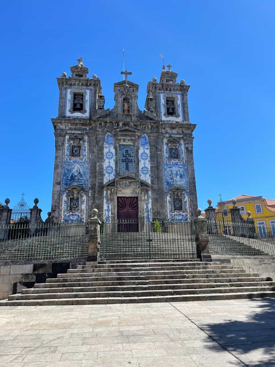 A beautiful view of the Church of Saint Ildefonso in Porto, featuring its striking blue and white azulejo tile facade. The baroque-style church stands at the top of a wide stone staircase, framed by two tall bell towers. The intricate tiles and grand entrance create a captivating scene against the clear blue sky.