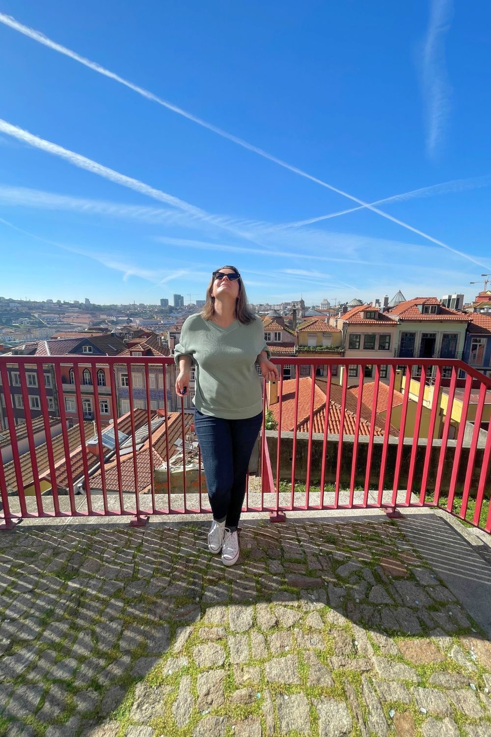 A woman on her own standing against a red fence with the city of porto in the background. 