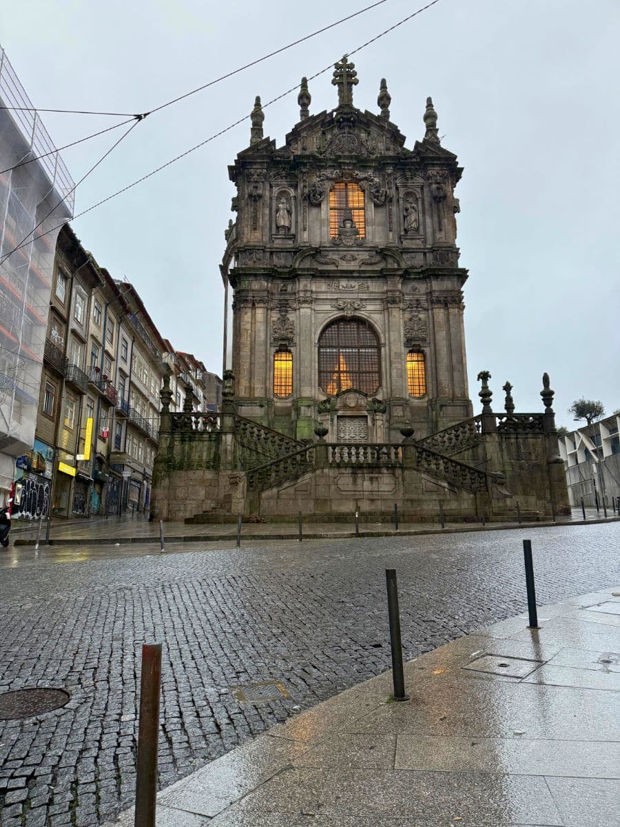 Rainy day view of the baroque Clérigos Church in Porto, showcasing its ornate façade with glowing windows, set against a backdrop of wet cobblestone streets and surrounding buildings.