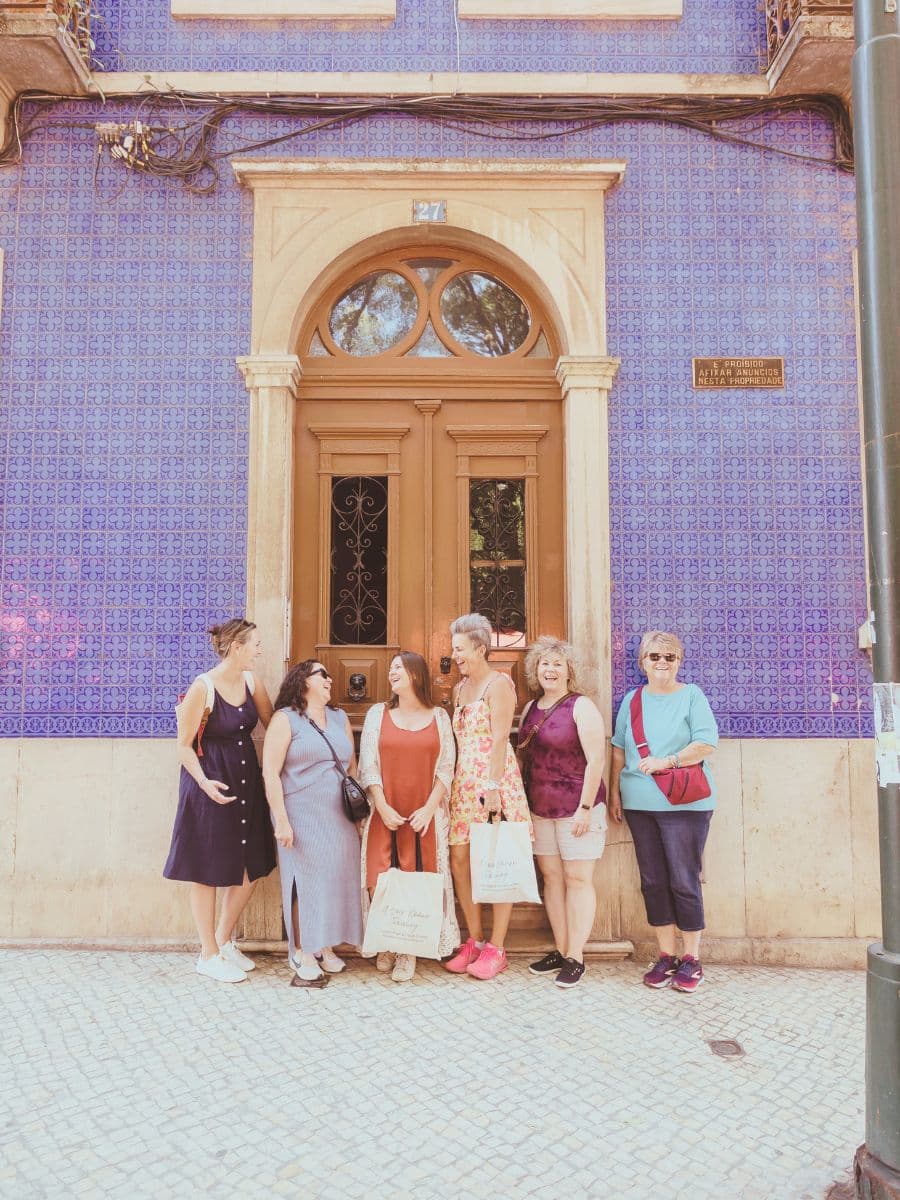 A joyful small group trip in Europe, featuring six women laughing together in front of a traditional blue-tiled building in Lisbon, Portugal. They are dressed casually, carrying tote bags, and enjoying a shared travel experience.