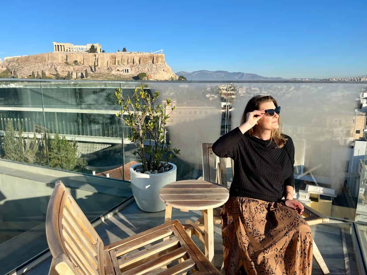 Woman sitting on a rooftop terrace in Athens, Greece, with a view of the Acropolis in the background, wearing sunglasses and a long skirt, looking up toward the sky.