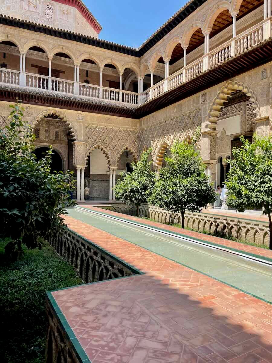 A photo of a courtyard inside the Real Alcázar in Seville, Spain. The courtyard features intricate Moorish arches, detailed carvings, a central reflecting pool, and lush orange trees, with the palace’s ornate upper balconies visible above.