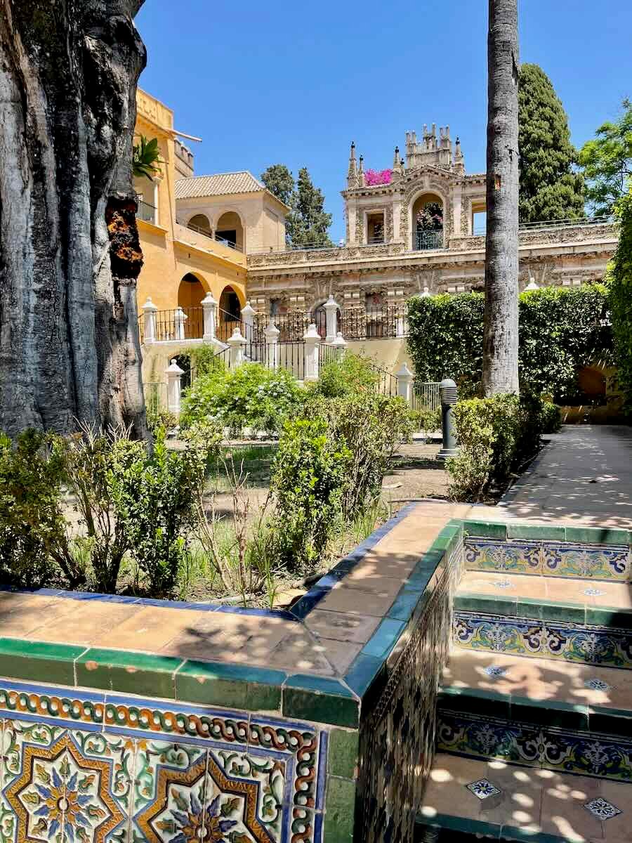 A photo taken in the gardens of the Real Alcázar in Seville, Spain. The image features a tiled staircase leading up to a garden area, with lush vegetation, tall trees, and ornate structures visible in the background.