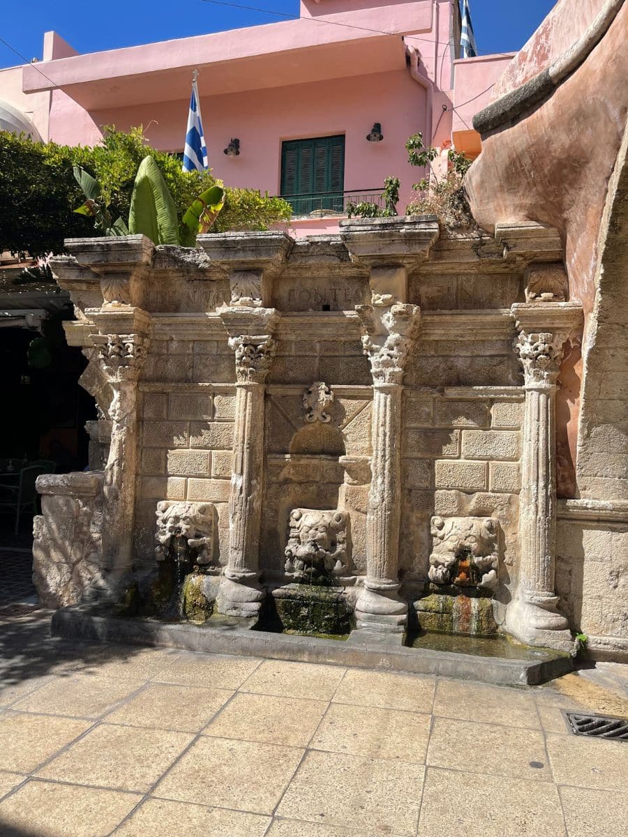 Ornate ancient Roman fountain adorned with detailed sculptures, nestled against a modern pink building in Crete, blending historical art with contemporary architecture.