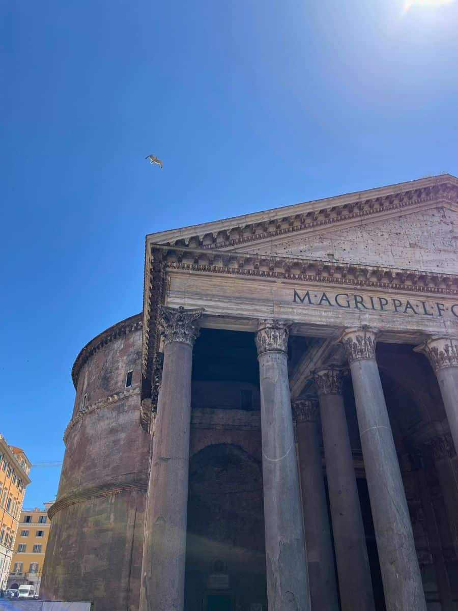 A partial view of the Pantheon in Rome, Italy, showcasing its grand Corinthian columns and the inscription on the pediment, 'M·AGRIPPA·L·F·COS·TERTIVM·FECIT.' The ancient Roman temple's distinctive rotunda is visible to the left, displaying its weathered brick exterior. A single seagull soars in the clear blue sky above, adding a dynamic element to the serene atmosphere. The sun casts strong shadows on the facade, highlighting the depth and texture of the Pantheon’s historic architecture.