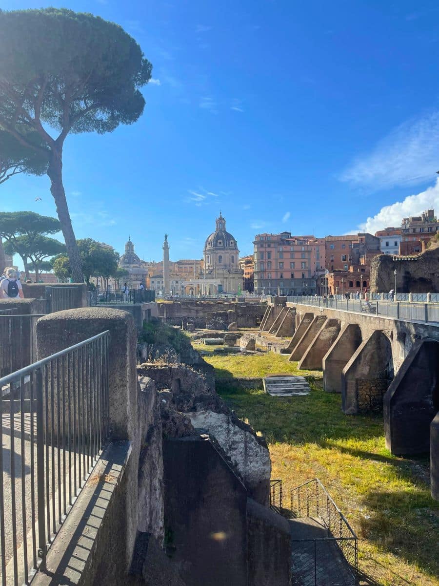 A scenic view of ancient ruins in Rome, Italy, with a tree-lined walkway on the left and historic buildings in the background. The Church of the Most Holy Name of Mary, with its distinctive dome, stands prominently in the distance alongside Trajan's Column. The ruins, bathed in sunlight, display remnants of stone walls and structures, offering a glimpse into Rome’s rich history. A bright blue sky with a few wispy clouds enhances the contrast between the old and new elements of the city.