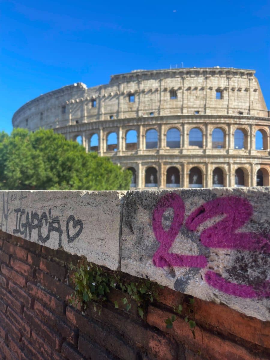 A view of the Colosseum in Rome, Italy, with a graffiti-marked stone and brick wall in the foreground. The graffiti includes a pink '23' and black lettering with a heart symbol. The ancient amphitheater, partially obscured by green trees, stands against a clear blue sky. The contrast between the historic architecture and modern graffiti highlights the coexistence of past and present in the city.