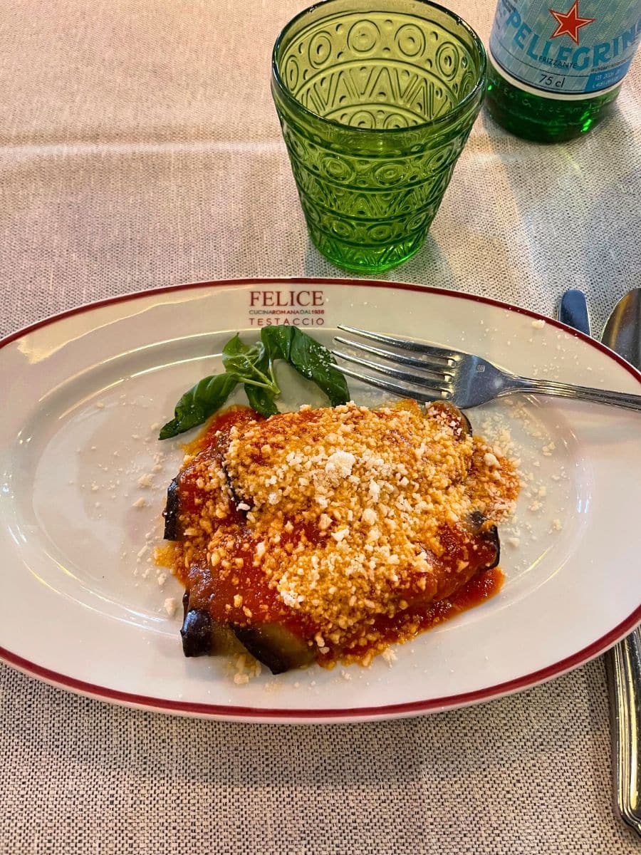 A plate of eggplant parmesan at Felice a Testaccio, a well-known restaurant in Rome, Italy. The dish is covered in tomato sauce and sprinkled with grated cheese. A basil garnish is on the side. A green glass and a bottle of San Pellegrino sparkling water are also visible on the table, set on a beige tablecloth