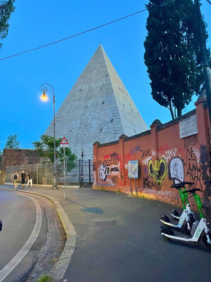The Pyramid of Cestius, an ancient Roman pyramid in Rome, Italy, at dusk. The white marble structure stands tall against the blue evening sky, surrounded by a black iron fence. A graffiti-covered red wall runs along the sidewalk, contrasting the historical monument with urban street art. Lime electric scooters are parked on the sidewalk, and a few pedestrians walk along the curved street.
