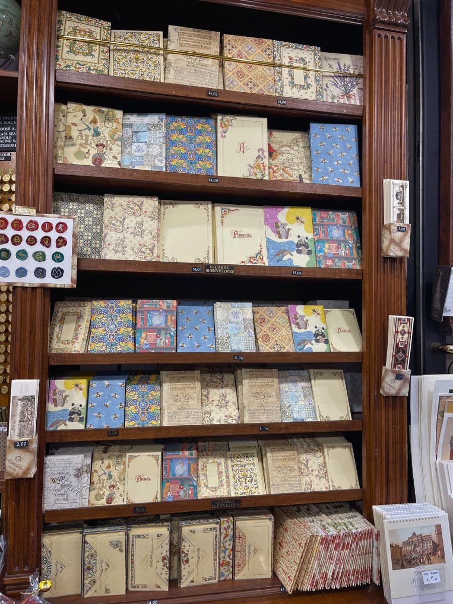 A display shelf in a stationery shop featuring an assortment of beautifully designed notebooks, journals, and writing sets with intricate Renaissance and floral-inspired patterns. The collection includes ornate gold-embossed designs, vintage-style illustrations, and decorative envelopes. The wooden shelving adds to the classic and elegant atmosphere, evoking a sense of old-world charm. Wax seals and postcards are also available, enhancing the store’s antique aesthetic.