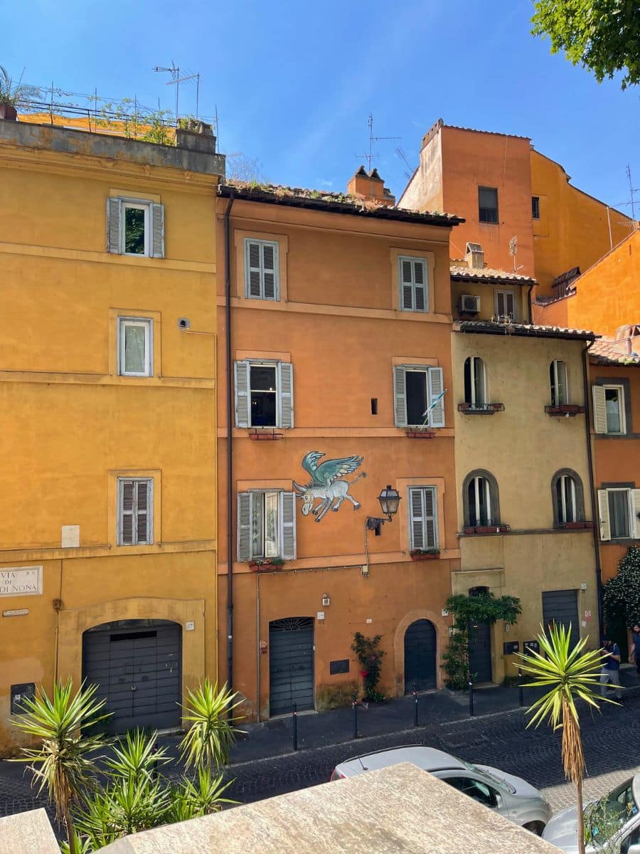 A charming street scene in Rome featuring warm-toned historic buildings painted in shades of ochre and terracotta. The architecture showcases traditional shuttered windows and arched doorways. A playful mural of a blue flying horse with wings adorns the façade of one building, adding a whimsical touch. Greenery, including potted plants and palm trees, lines the cobblestone street, enhancing the Mediterranean atmosphere under a bright blue sky.