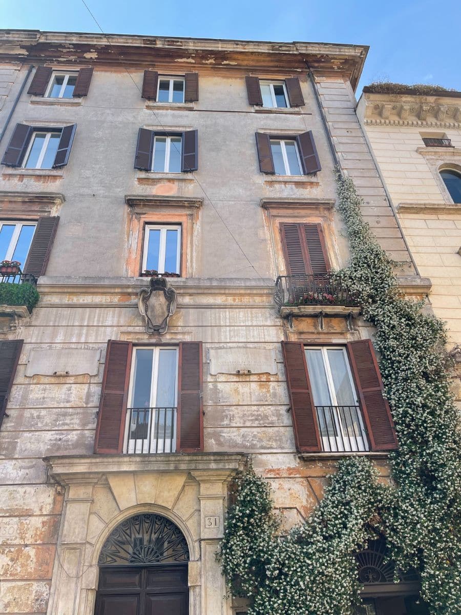 A charming old European building with a weathered facade and rustic wooden shutters. Climbing ivy with delicate white flowers partially covers the lower part of the structure. The building features tall windows, some with wrought iron railings, and an ornate arched entrance with intricate detailing above the dark wooden door. The warm tones of the peeling plaster contrast beautifully with the blue sky above.