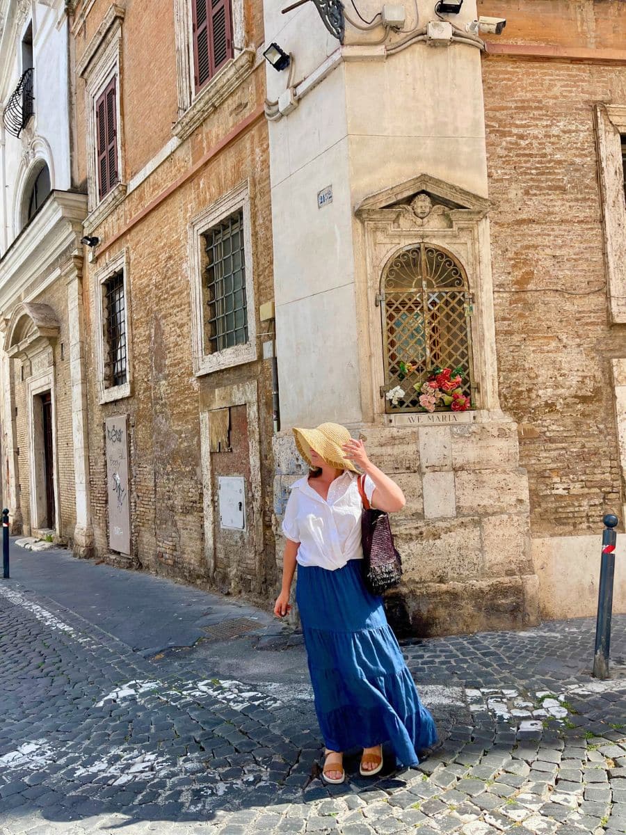 A woman dressed in a white blouse, a flowing blue skirt, and a wide-brimmed straw hat walks along a cobblestone street in Rome, Italy. She shields her face from the sun with her hand while carrying a woven bag over her shoulder. The background showcases a historic brick and stone building with aged, textured walls and a small religious shrine embedded in the corner, adorned with flowers. The scene is illuminated by bright sunlight, casting strong shadows and emphasizing the warm tones of the architecture.