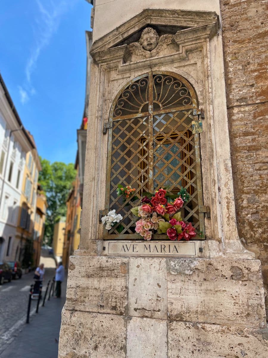 A close-up of a small religious shrine embedded in the corner of an aged stone building in Rome, Italy. The shrine features an arched metal grate with intricate scrollwork, adorned with a bouquet of artificial red, pink, and white flowers. Above the grate, a sculpted cherub's face with wings is carved into the stone. Below the shrine, a plaque with the inscription 'AVE MARIA' is embedded in the weathered stone, reflecting the religious devotion of the site. In the background, a narrow cobblestone street lined with historic buildings and a few pedestrians is visible under a bright blue sky.