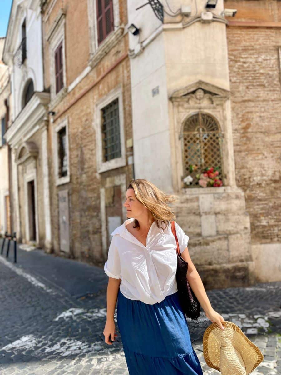 Melissa with shoulder-length brown hair walks down a cobblestone street in Rome, Italy, bathed in warm sunlight. She wears a flowing white button-up shirt and a long blue skirt, carrying a woven straw hat in one hand and a black tote bag over her shoulder. Her gaze is directed to the side, with the breeze gently tousling her hair. Behind her, an old brick building with a small religious shrine featuring an ornate metal grate, flowers, and an 'Ave Maria' plaque is visible. The surrounding buildings have rustic charm with aged facades, wooden shutters, and traditional Roman architecture.