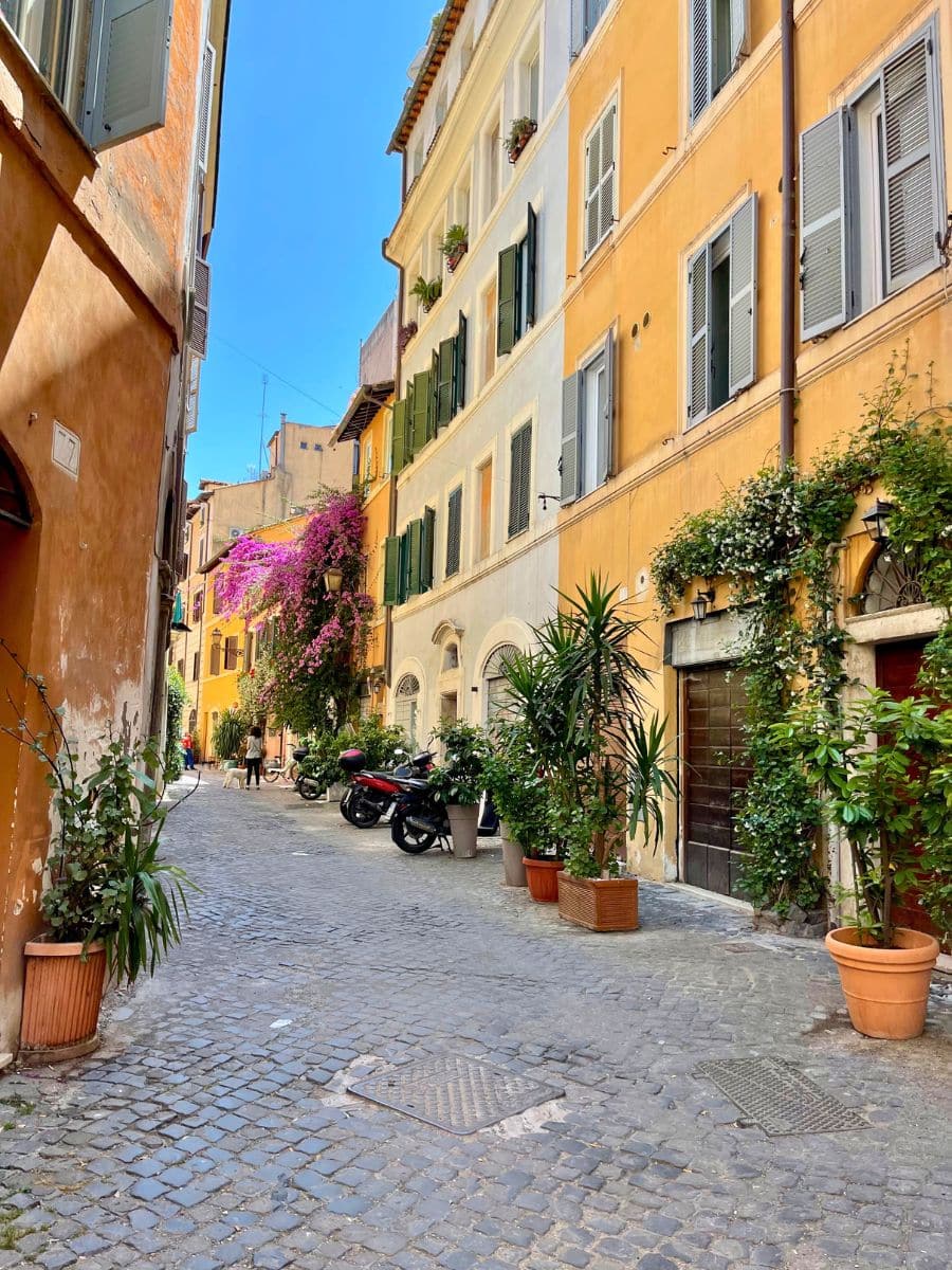 A picturesque narrow street in Rome, Italy, paved with cobblestones and lined with colorful historic buildings in warm shades of yellow and ochre. Green wooden shutters and potted plants decorate the facades, while vibrant pink bougainvillea cascades from a balcony. Motorbikes are parked along the street, and a few pedestrians stroll in the distance, adding to the charming, lively atmosphere of this quaint Roman alleyway bathed in sunlight.
