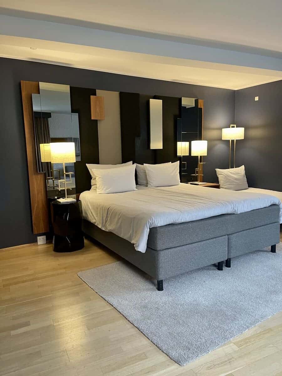 Elegantly appointed bedroom at Hotel Skt Petri with a large gray bed, multiple decorative mirrors and lamps, dark paneling creating a contemporary look, and a soft gray rug, combining comfort and style.