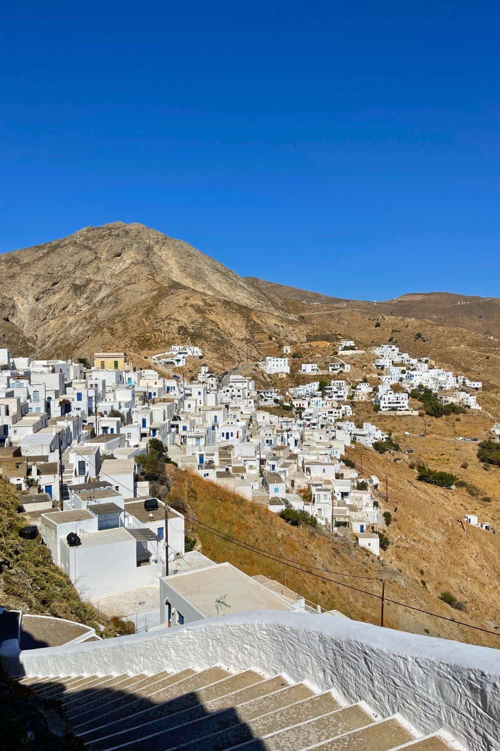 The image shows a cluster of whitewashed buildings on a hillside under a clear blue sky. The buildings are densely packed, creating a striking contrast against the dry, rocky terrain surrounding them. In the background, a barren mountain rises, adding to the rugged beauty of the scene. The stairway in the foreground hints at the steep and winding paths through the village.