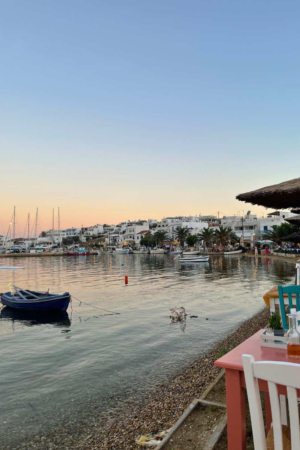 The image shows a peaceful harbor at sunset, with calm waters reflecting the soft evening light. Small boats are docked along the shore, and buildings are visible in the distance. The foreground includes a seating area near the water, adding a cozy, relaxed feel to the scene. The sky fades from light blue to soft pink, enhancing the tranquil atmosphere.