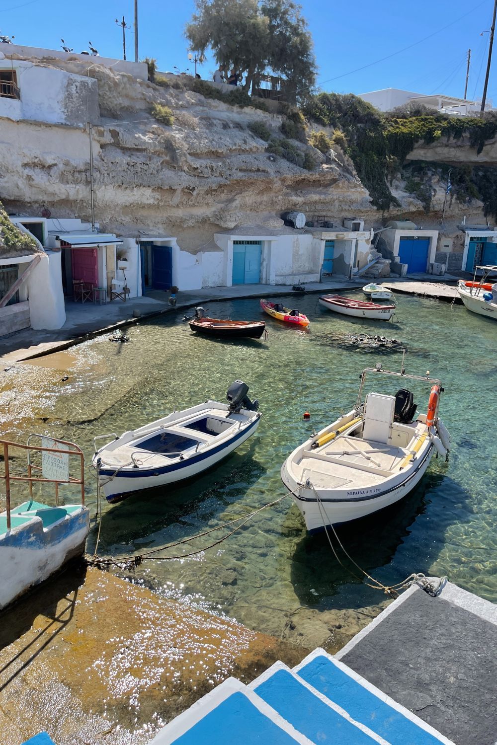 Colorful boats floating in clear water by a rugged cliff with traditional white and blue fishermen's houses in Milos.