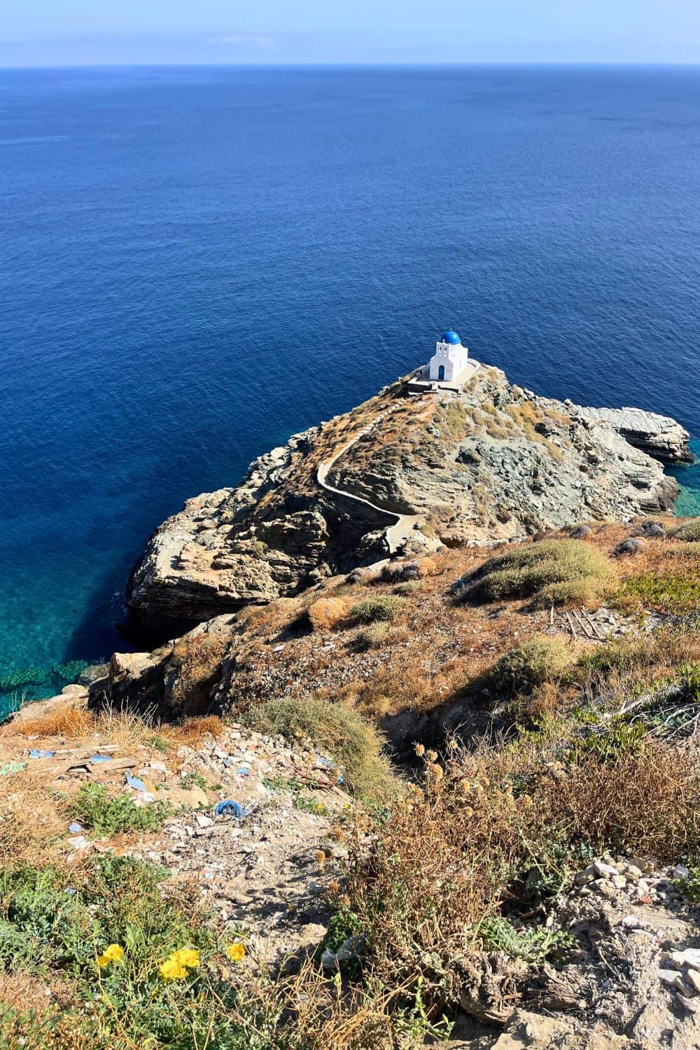 The image shows a serene coastal scene with a small white chapel or structure situated on a rocky peninsula that juts out into a calm, deep blue sea. The rocky terrain surrounding the structure is dotted with low vegetation, giving it a rugged but peaceful appearance. A narrow pathway leads up to the chapel, creating a sense of isolation and tranquility. The horizon stretches into the distance, blending the sea and sky. This image likely depicts a remote and picturesque location, possibly along the Mediterranean coast.