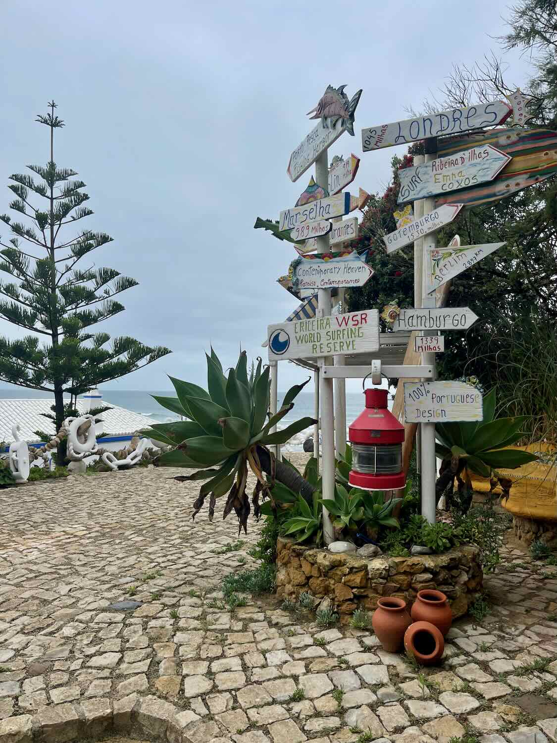 A whimsical outdoor scene in Ericeira, Portugal, featuring a signpost with multiple colorful, hand-painted directional signs pointing to various destinations. The signpost is surrounded by lush greenery, including succulents and agave plants, set on a cobblestone patio. In the background, a view of the ocean is partially visible under an overcast sky.