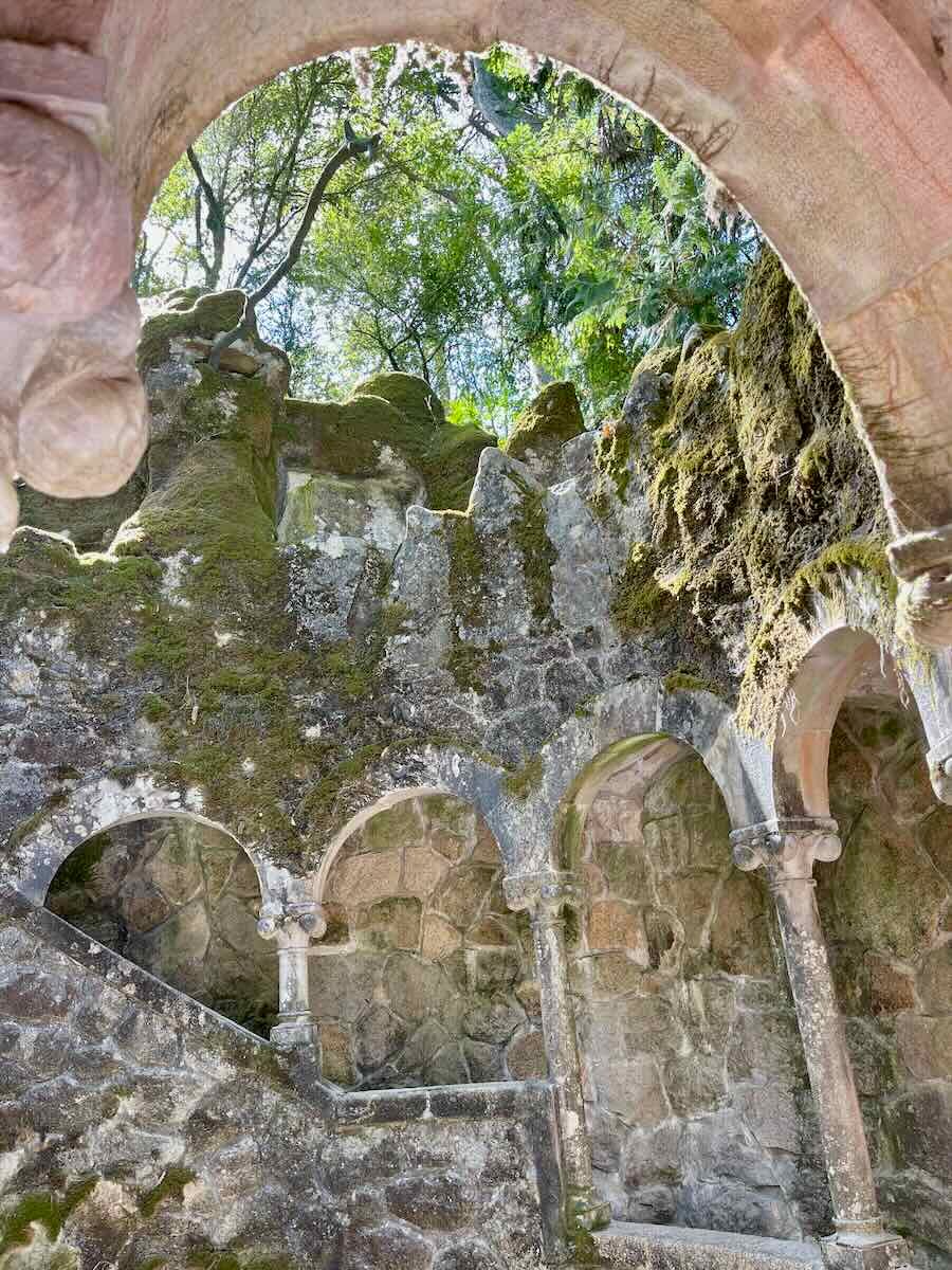 An ancient stone staircase winds through a moss-covered, rocky structure with several archways. Sunlight filters through the trees above, creating a serene and mystical atmosphere in this hidden corner of Sintra.