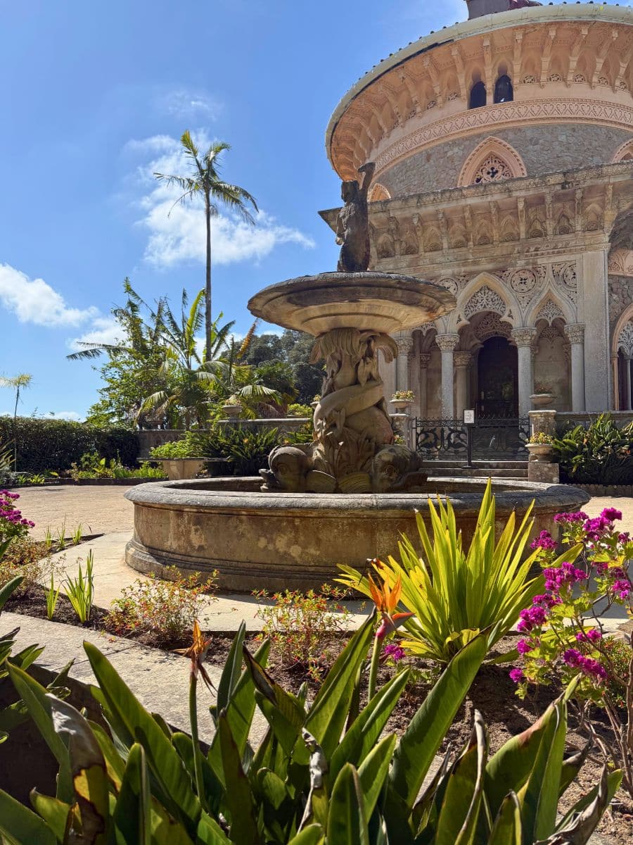 Ornate round palace facade in Sintra with arched windows and detailed stone carvings, framed by a decorative fountain in the foreground and vibrant tropical plants under a bright blue sky.