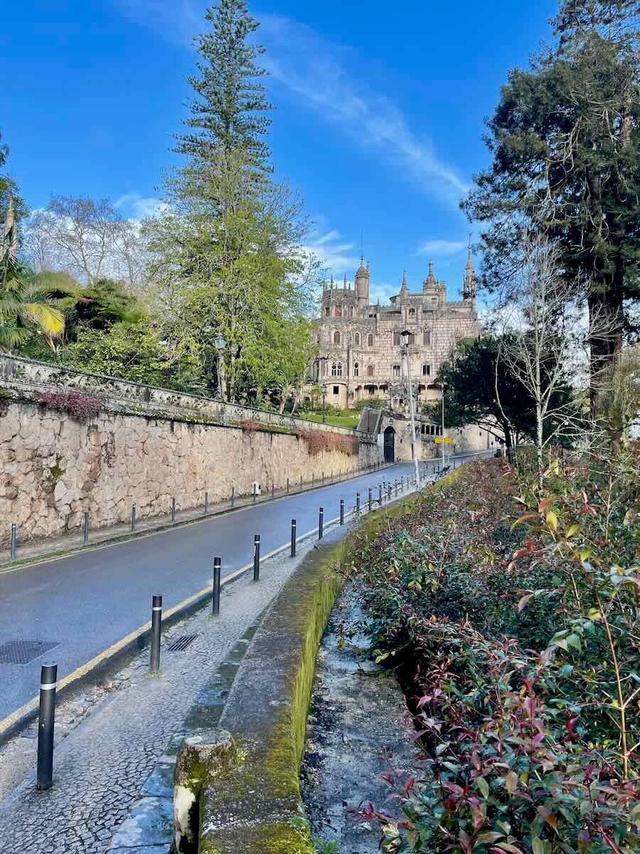 A picturesque view of a stately palace in Sintra, Portugal, with its gothic architecture seen from a quiet road lined with trees and shrubs. The road curves gently, leading toward the majestic structure framed by greenery.