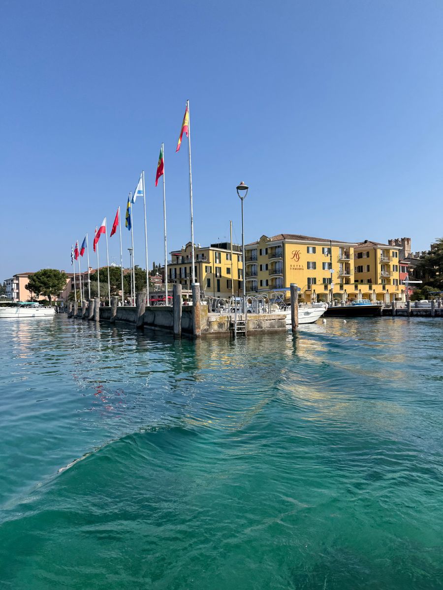 A vibrant waterfront scene in Sirmione, Italy, featuring turquoise waters gently rippling in the foreground. A row of flagpoles displaying various national flags lines the edge of a small harbor. Behind the flags, the yellow facade of Hotel Sirmione e Promessi Sposi stands prominently under a clear blue sky. Boats are docked along the pier, and the charming architecture and serene setting suggest a peaceful, sunny day on Lake Garda.